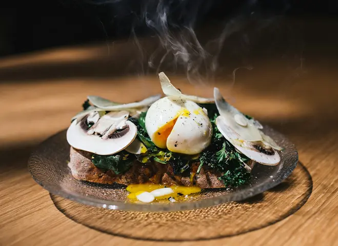 a plate with a variety of thinly-sliced cooked mushrooms with a boiled egg and some greens on a piece of toast on a small glass plate.